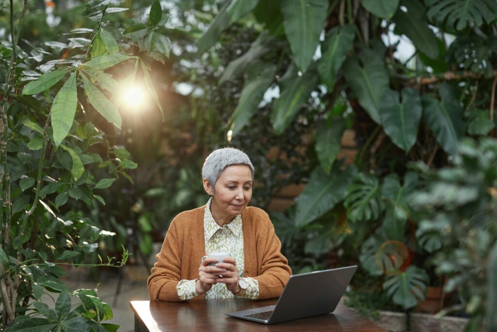 Senior woman enjoys coffee and laptop in a tropical garden, embracing nature and technology.