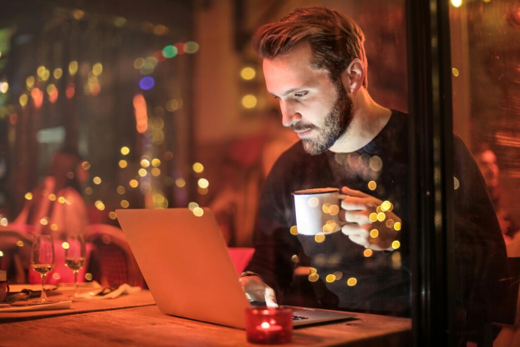 A young man with a beard is working on a laptop at a cozy cafe with warm lighting, holding a coffee cup.